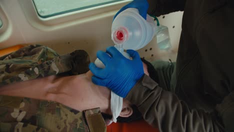 close up of a confident male army medic in a dark green uniform and blue gloves doing artificial respiration to an unconscious man using a matte white special bottle for artificial respiration in the ambulance cabin