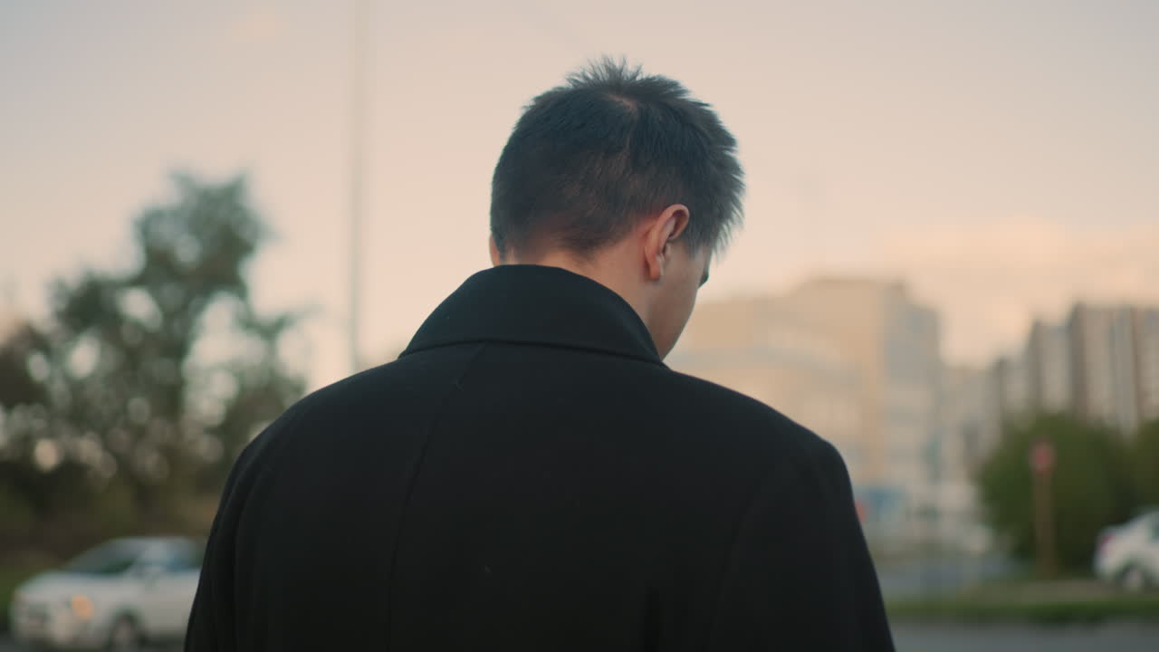 검은 코트를 입은 개인이 차를 마시며 도시 환경에서 사려 깊게 앞을 바라보고있는 뒷면, 부드러운 빛 아래 자동차와 높은 건물로 흐릿한 배경을 특징으로합니다.