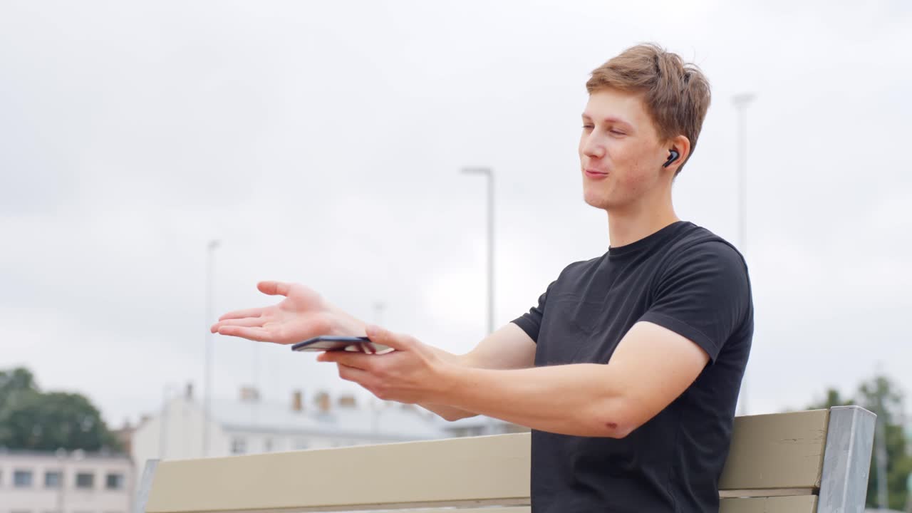 Young man using a smartphone and wireless earbuds for a video call outdoors