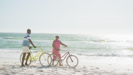 Happy-senior-african-american-couple-walking-with-bikes-at-beach-with-copy-space,-in-slow-motion