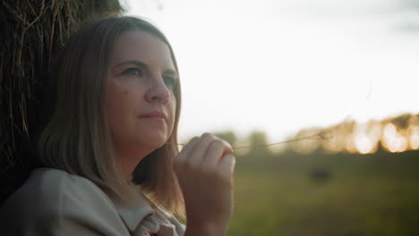 close-up of woman in deep thought holding hay strand, gazing into distance, warm golden light enhances blurred background, creating a serene and reflective