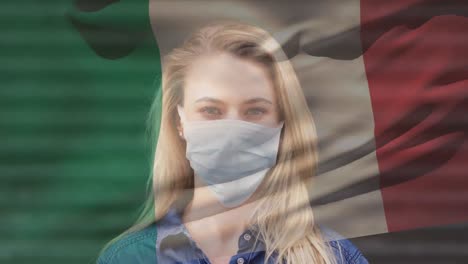 animation of flag of italy waving over woman wearing face mask during covid 19 pandemic