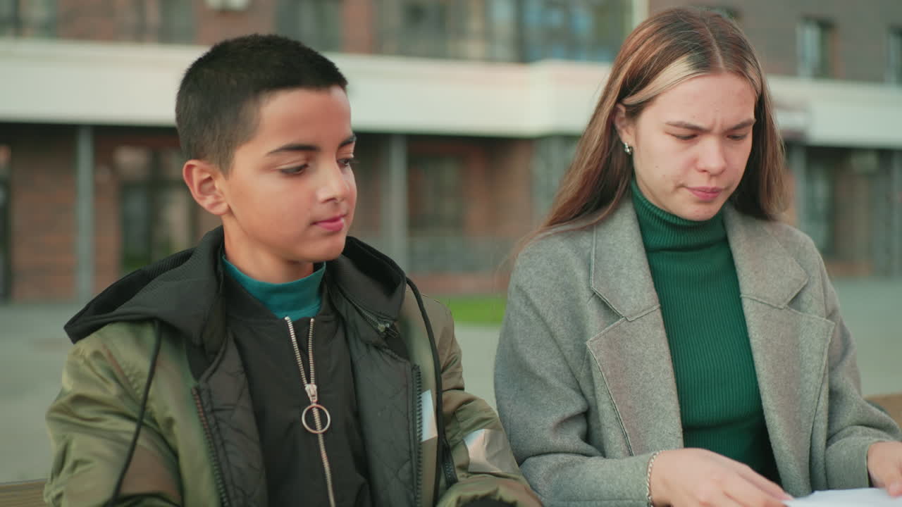 Boy looks at lady smiling as she folds paper while sitting outdoors at wooden table, both engaged in creative activity, showing focus, learning, and joyful expression in relaxed urban environment