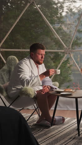 couple has breakfast with coffee and pancakes on glamping terrace. woman and man in bathrobes spend morning sitting at table with view on lake and mountains