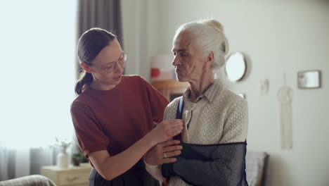 young woman helping elderly grandmother adjust arm sling at home