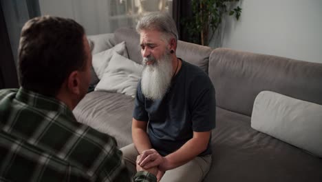 Over-the-shoulder-A-sad-man-with-gray-hair-and-a-lush-gray-beard-in-a-gray-T-shirt-communicates-with-his-boyfriend-a-brunette-with-stubble-in-a-checkered-shirt-and-holds-his-hands-while-sitting-on-a-gray-sofa-during-a-difficult-conversation-among-men-in-an-LGBT-couple-in-the-evening-in-a-modern-apar