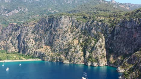 aerial drone panning up towards the large green mountains of butterfly valley while boats are anchored in the blue ocean of fethiye turkey