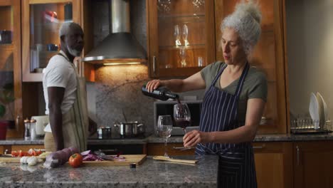 Caucasian-senior-woman-wearing-apron-pouring-wine-in-the-glass-in-the-kitchen-at-home