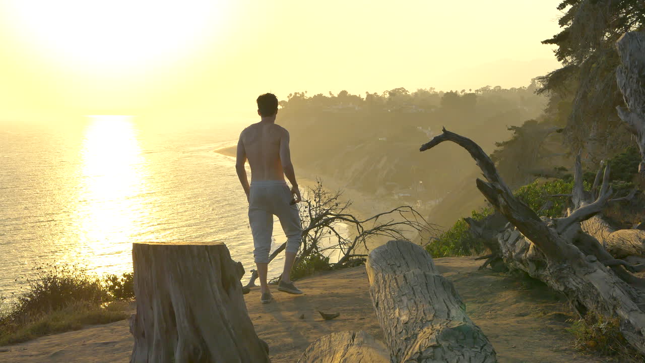 Jovem em forma, sem camisa, caminhando até a beira de um penhasco após uma corrida acima do Oceano Pacífico durante um pôr do sol dourado em Santa Bárbara, Califórnia.