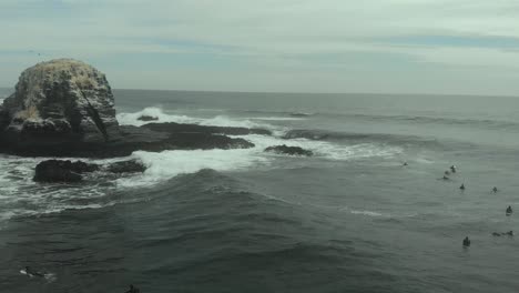 Aerial-orbit-and-reveal-of-rocky-formations-and-surfers-waiting-for-a-wave-in-Pichilemu,-Chile-4K