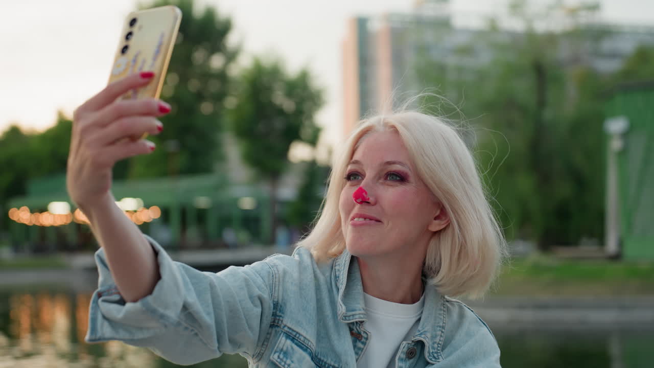 young woman with painted nose holding smartphone at arm length for selfie by river park at golden hour during picnic, denim jacket, smiling, blurred trees and lights reflecting on water