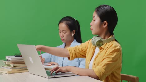 close up of asian woman student typing on a laptop and talking on smartphone while sitting with her friend studying on a table in the green screen background classroom