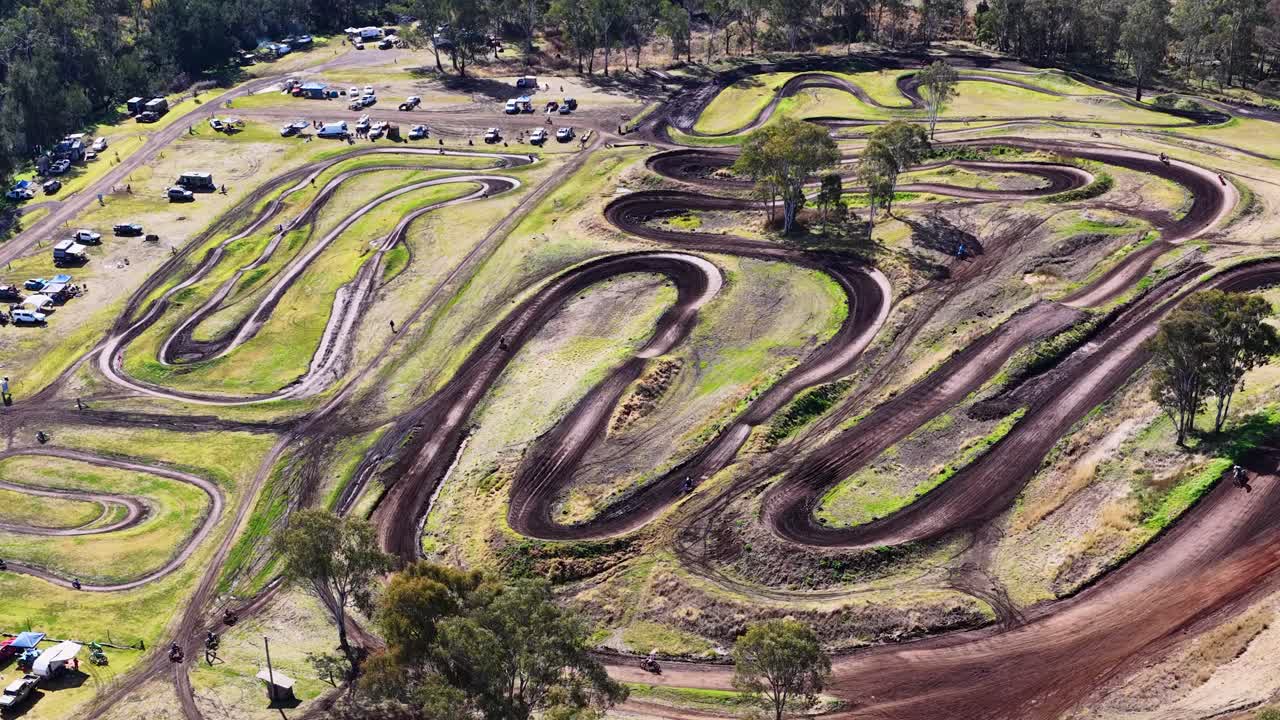 Drone footage captures motorbikes racing and jumping on a winding dirt track surrounded by bushland, vehicles, and camping areas in bright daylight