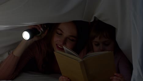 young mother reading goodnight story fairytale to child daughter under duvet blanket in night room