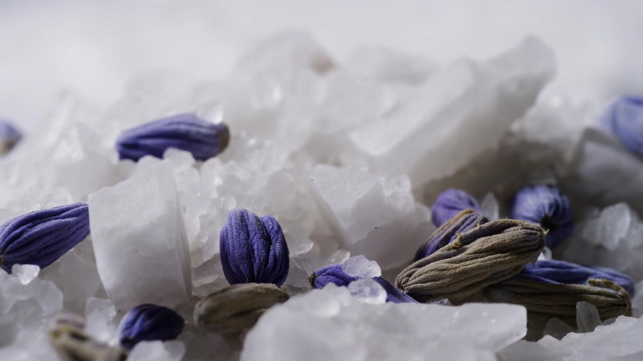 Scena dettagliata di boccioli di lavanda essiccati annidati tra cristalli di sale traslucidi e irregolari, con un sottile assestamento nelle fessure sotto una luce morbida e diffusa, creando una texture naturale e calma e un'atmosfera da spa rilassante.