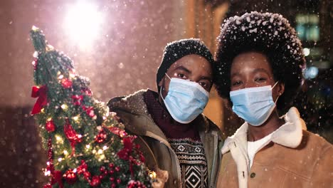 close-up view of joyful african american couple wearing facial masks holding a christmas tree and smiling at camera while it¬¥s snowing on the street in christmas