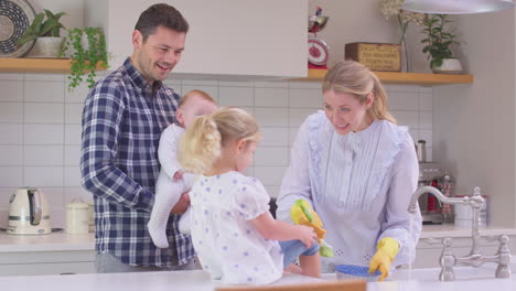 Vater-Und-Kleiner-Sohn-Schauen-Zu,-Wie-Die-Tochter-Auf-Der-Theke-Sitzt-Und-Der-Mutter-Beim-Abwaschen-Des-Geschirrs-Im-Waschbecken-Hilft-–-Aufgenommen-In-Zeitlupe