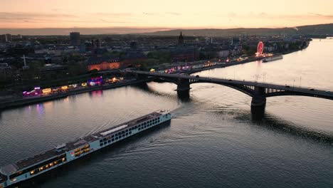 Vuelo-De-Drones-Al-Atardecer-De-Mainz-Sobre-El-Río-Rin-Cruzando-Un-Barco-Y-El-Puente-Principal-De-La-Ciudad-Frente-Al-Cielo-Naranja