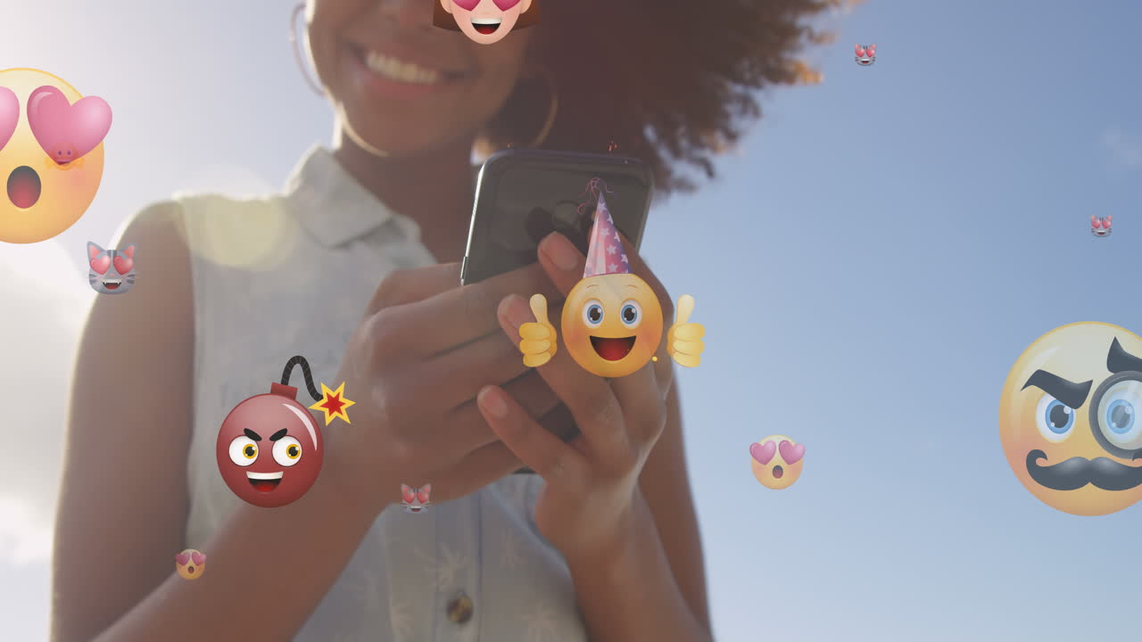 animación de emoticones sobre la sección media de una mujer biracial que usa un teléfono inteligente al aire libre