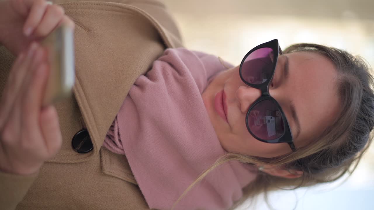 Brunette woman wearing a brown coat scrolling on her phone at a terrace. Vertical