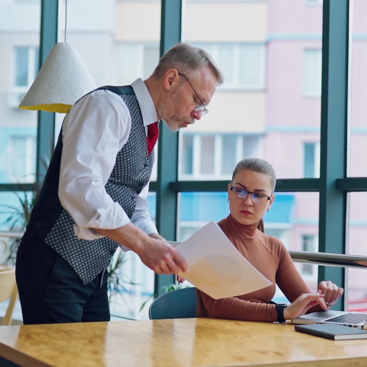 Young woman working on a computer near the window. Senior businessman coming to his secretary sitting at the desk and showing her some papers. Slow motion.