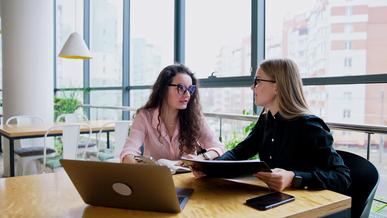 Young attractive women having conversation at office. Two ladies sit at desk having phone, documents and laptop in front of them.