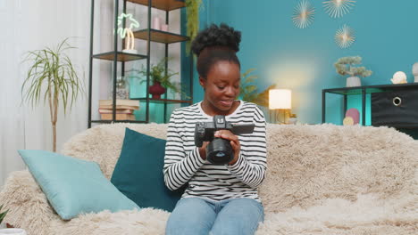 african american smiling young woman while reviewing successful photos on camera sits on home sofa