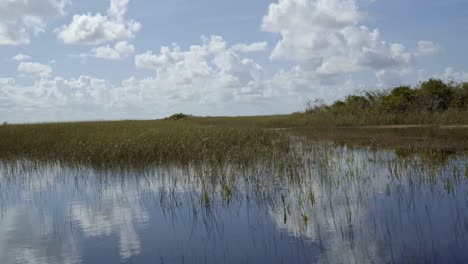 Schnelles-Trucking,-Linke-Aufnahme-Der-Atemberaubenden-Florida-Everglades-In-Der-Nähe-Von-Miami,-Fahrt-Auf-Einem-Luftkissenboot,-Während-Das-Ruhige-Sumpfwasser-Den-Himmel-Reflektiert-Und-An-Einem-Sonnigen-Tag-Eine-Fata-Morgana-Inmitten-Von-Hohem-Gras-Erzeugt