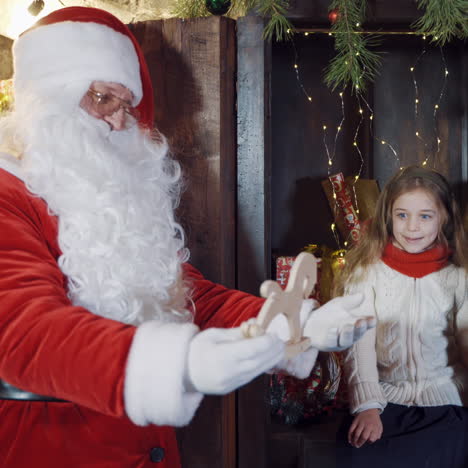 santa claus with a little girl. santa in red costume hold a wooden toy horse and gives it to a small girl indoors.