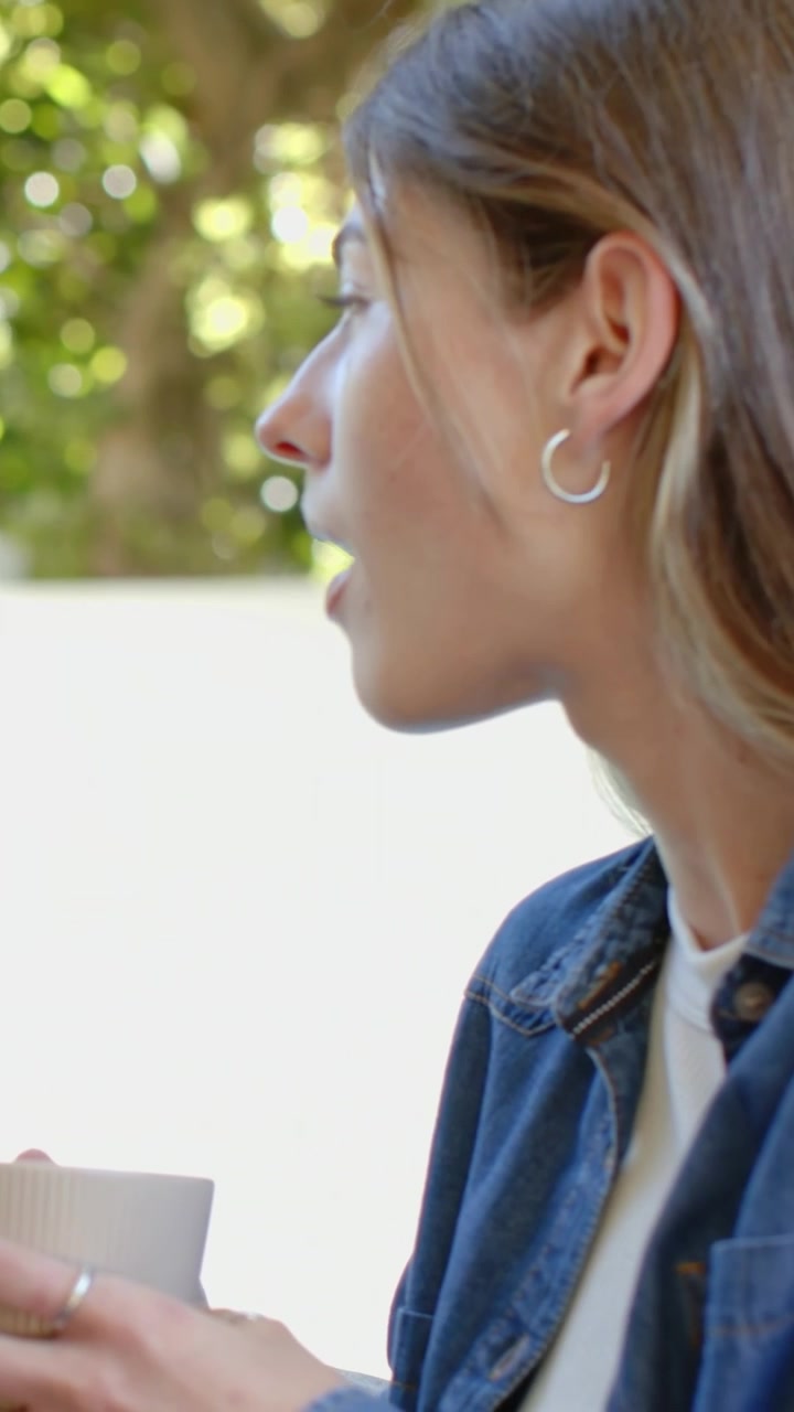 Video verticale: Donna in jeans che parla, tenendo una tazza sul patio, girandosi verso una chiamata fuori campo, con spazio per il testo