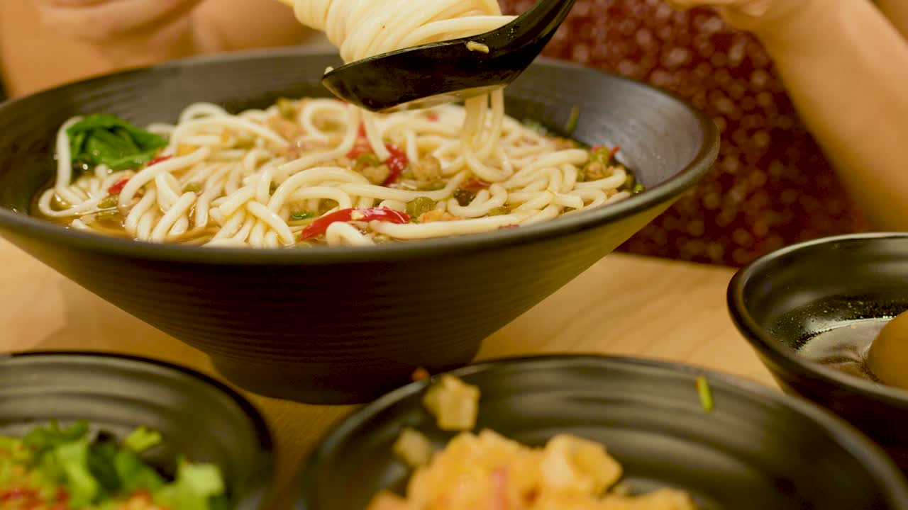 Woman uses chopsticks and spoon to eat spicy noodle soup at restaurant table, warm lighting