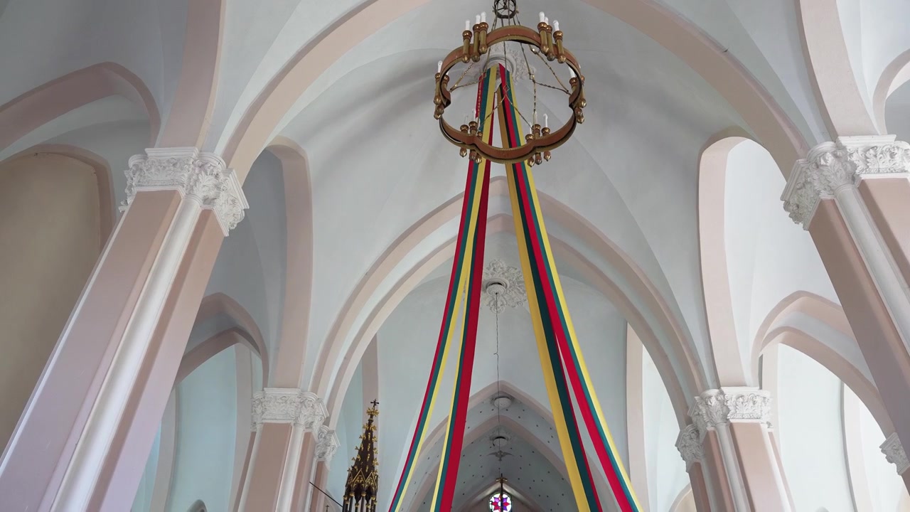 Ampia vista di un interno di chiesa luminoso e sfarzoso, adornato con lunghi nastri gialli, verdi e rossi sotto un grande lampadario, che evidenzia drammatici archi gotici e dettagli decorativi