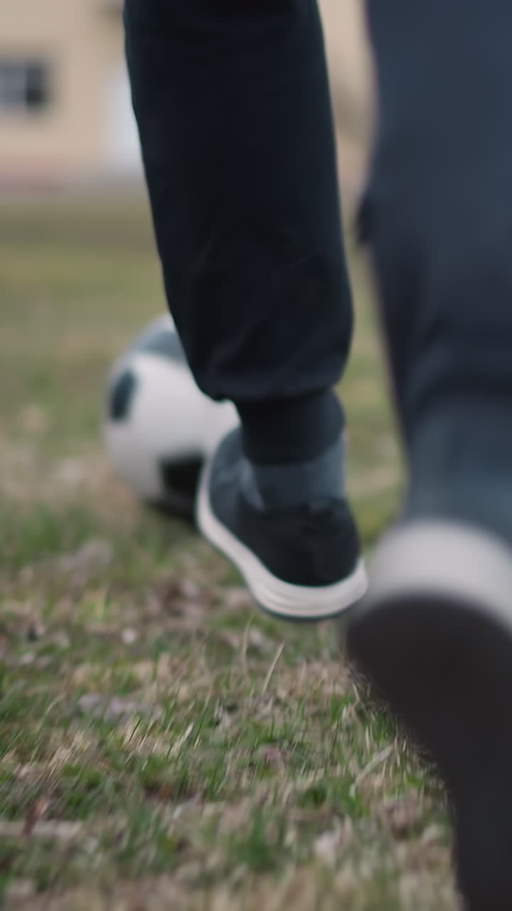 Close-up of someone in black pants and sneakers running with a soccer ball towards a goal post, with a blurred view of a person standing at the goal and buildings in the background