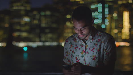 Mann-Benutzt-Nachts-Sein-Mobiltelefon-Mit-Der-Skyline-Der-Stadt-Im-Hintergrund