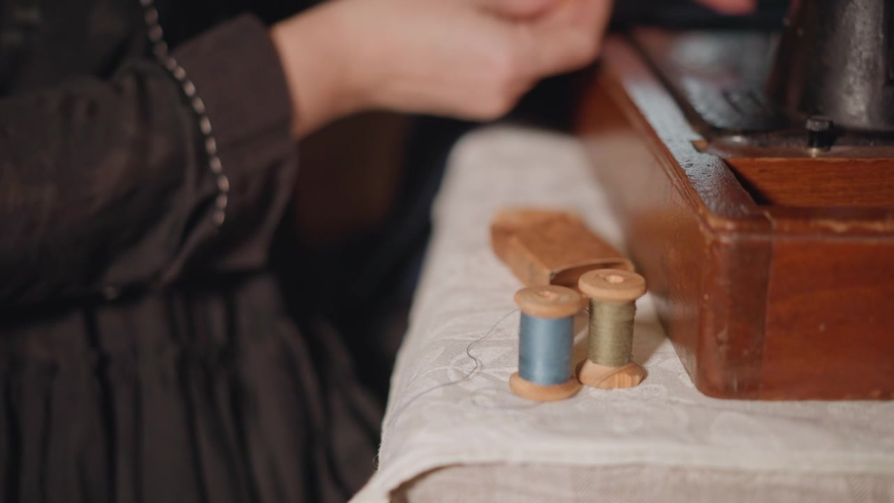 Rocchetti di filo accanto a una macchina da cucire antica, mani caucasiche che selezionano rocchetti blu sul tavolo, luce calda di una lampada, forniture organizzate e un tranquillo momento di artigianato domestico incentrato su colore e ordine.