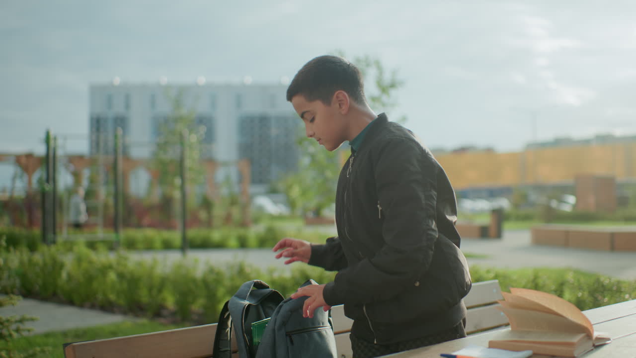 High school boy standing outdoors near bench packing study materials into backpack after completing study session, open book and notebook remain on wooden table