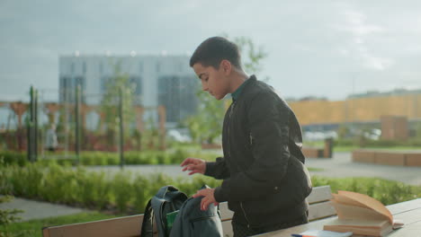 high school boy standing outdoors near bench packing study materials into backpack after completing study session, open book and notebook remain on wooden table
