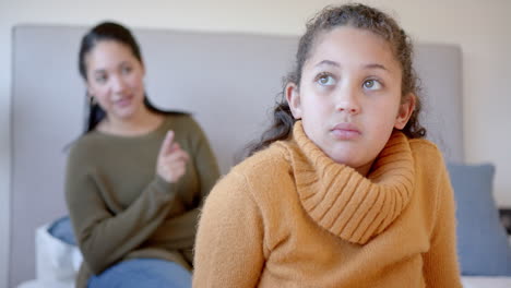 Nervous-biracial-mother-admonishing-daughter-on-bed-in-sunny-bedroom