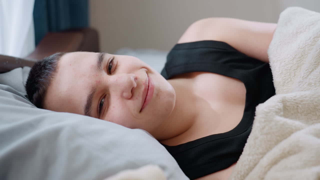 Close up boy lying on bed in black singlet opening eyes with gentle warm smile covered with blanket resting on pillow in cozy bright bedroom showing comfort