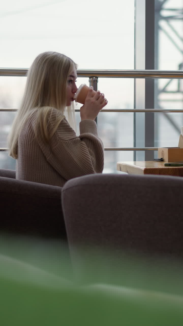 Sidebillede af ung kvinde, der holder en kaffekop og nipper til en drink i en hyggelig café i et indkøbscenter med sløret bybillede gennem vinduet, mens hun nyder et stille øjeblik med bystemning.
