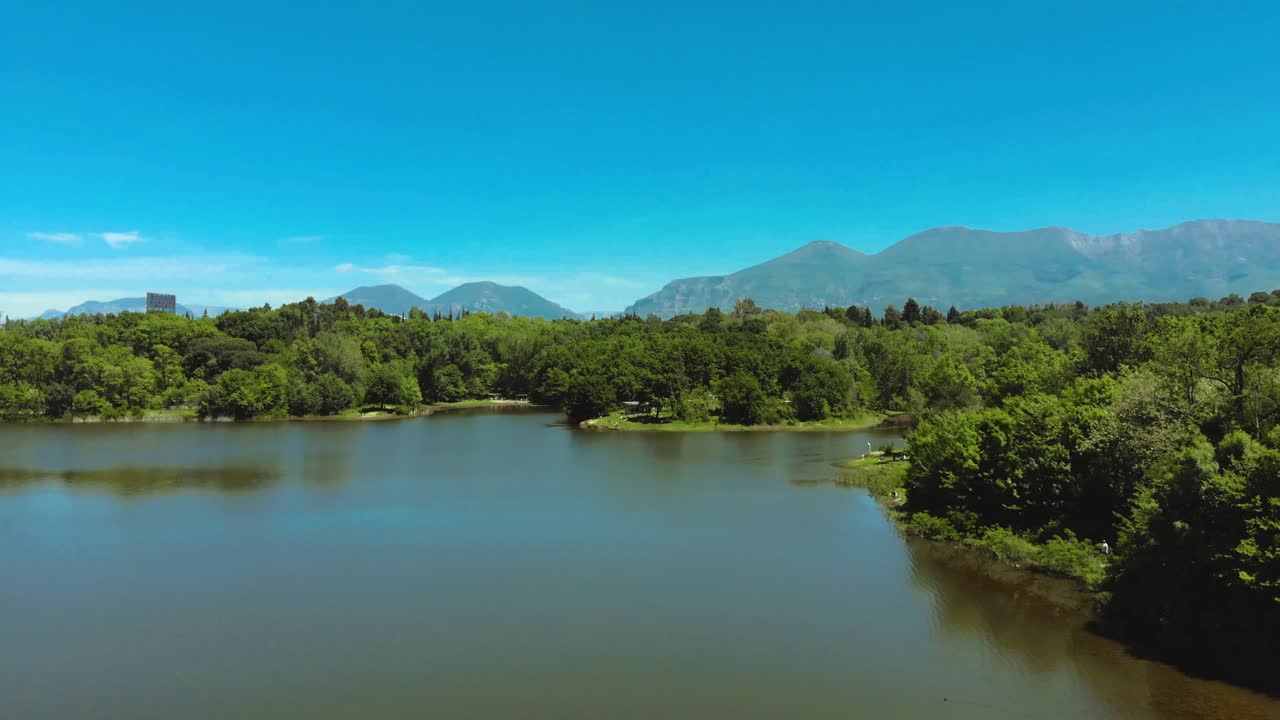 Green trees forest and meadows on park surrounded by calm lake water on a bright blue sky background