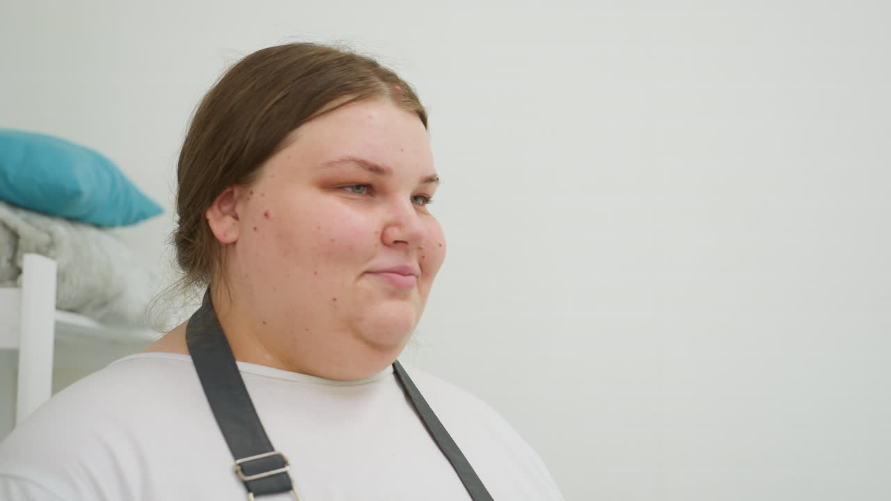 Beautician wearing white shirt and dark apron stands calmly in white room, positioned near stacked pillows and folded blanket on shelf in a clean salon environment with soft lighting