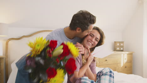 pareja con pijama en la cama en casa celebrando cumpleaños o aniversario con tarjetas y flores