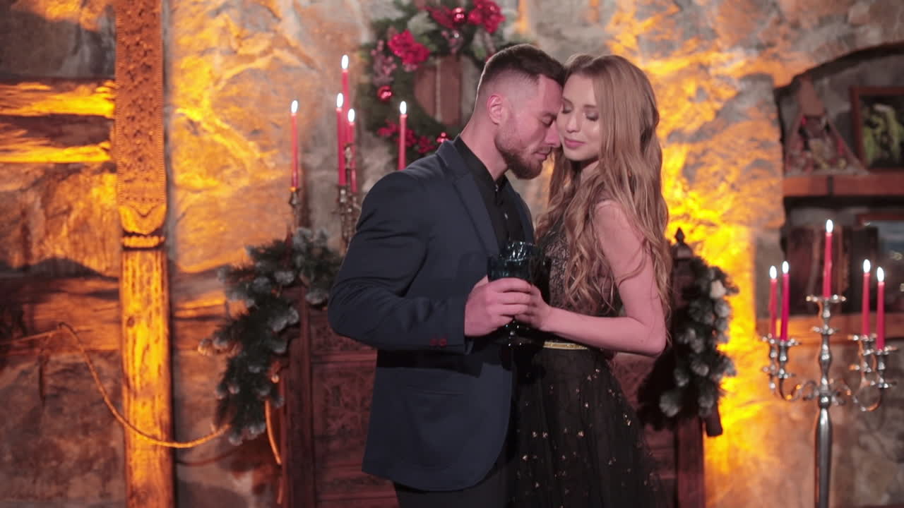 Embracing couple in beautiful clothes holding glasses. Happy pair in the room with garlands at Christmas season.