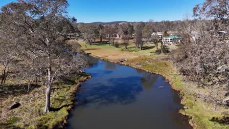 drone footage glides above a winding stream bordered by trees and grassy banks, revealing local residential houses in a quiet australian town under clear daylight