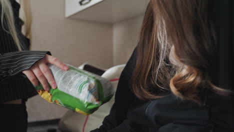 close up of sister seated on kitchen cabinet receiving bread and snacks from friend just returned from journey as she drops bag on cabinet and hands over treats with warm smiling interaction