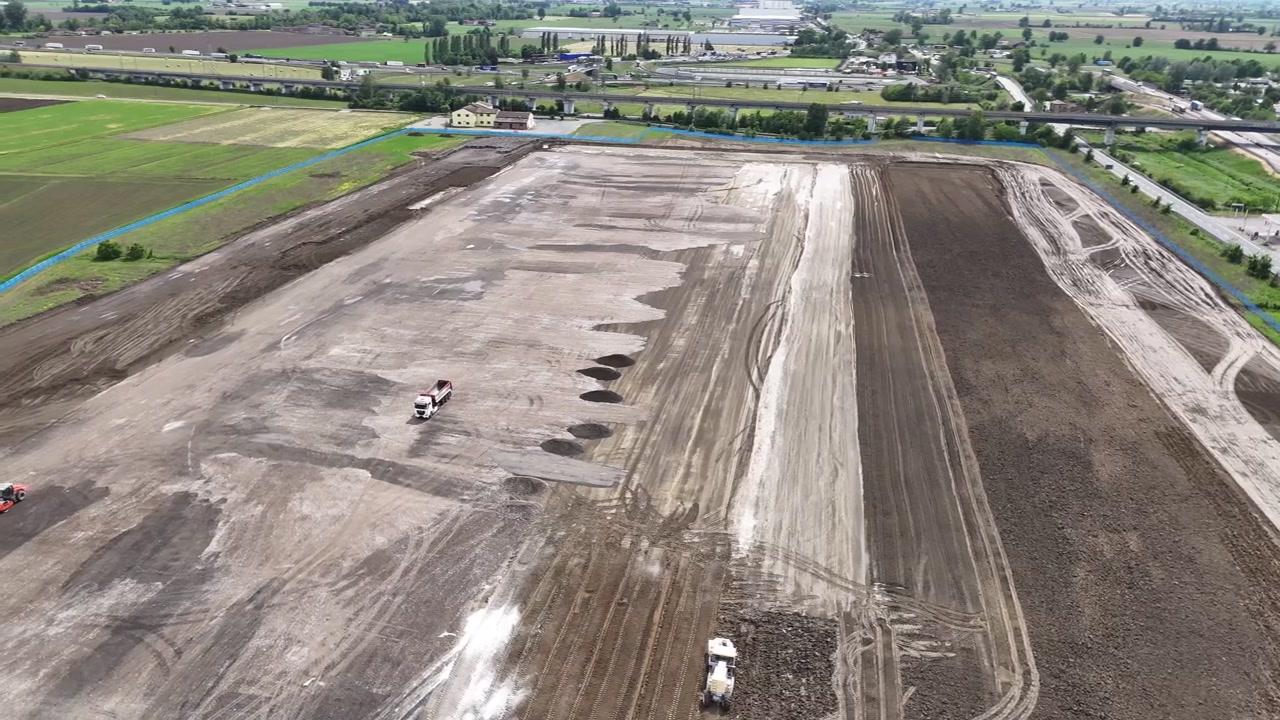 Attività di preparazione e compattazione del terreno su larga scala vicino all'infrastruttura di uno svincolo autostradale nella Pianura Padana, Italia, con corridoi livellati, macchinari pesanti e sviluppo di trasporti ingegnerizzati, vista aerea.