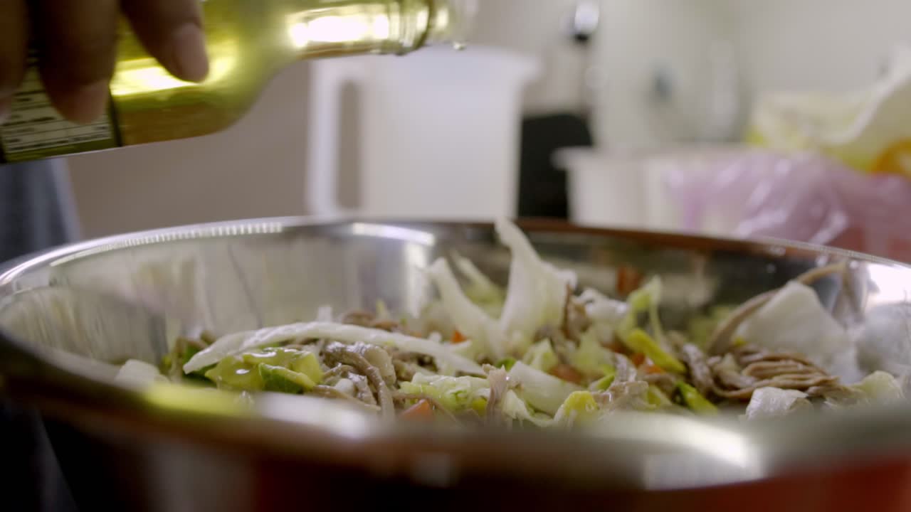 Close up of a chef pouring vegetable oil or dressing into a large stainless steel bowl filled with salpicon salad, featuring shredded meat and fresh vegetables