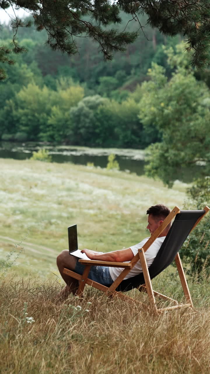 Freelancer works in a beautiful green place away from the city. Man sits in a folding chair with laptop on his knees. Vertical video