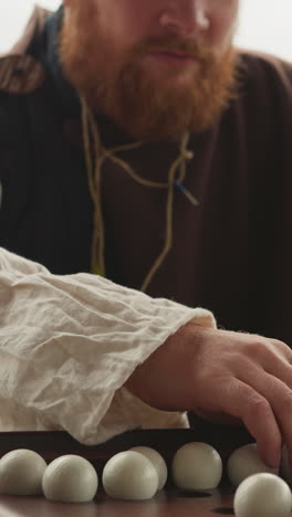 red-haired bearded man plays board game suspense badaboum. player in medieval clothes sits at table looking at opponent waiting for move closeup on blurred background